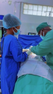 A doctor and a nursing student perform a procedure on a patient while wearing scrubs, face masks, and hair coverings.