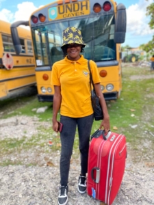A young Haitian woman wearing a yellow polo shirt and holding a red suitcase stands in front of a school bus and smiles. She also wears a black bucket hat printed with yellow flowers.