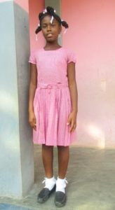 A young Haitian girl stands in front of a wall. She wears a pink dress, white socks, black loafers, and ribbons in her hair.