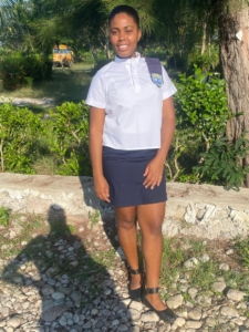 A young Haitian woman wearing a white blouse and a navy skirt stands in front of a tree and smiles. In the background is a school bus.