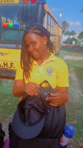 A young Haitian woman in a yellow polo shirt stands in front of a school bus and smiles. She holds a black backpack and a black baseball cap.