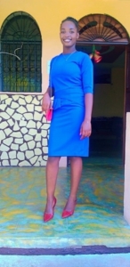 A young Haitian woman wearing a blue dress and red high heels and carrying a red purse stands in front of a doorway and smiles.
